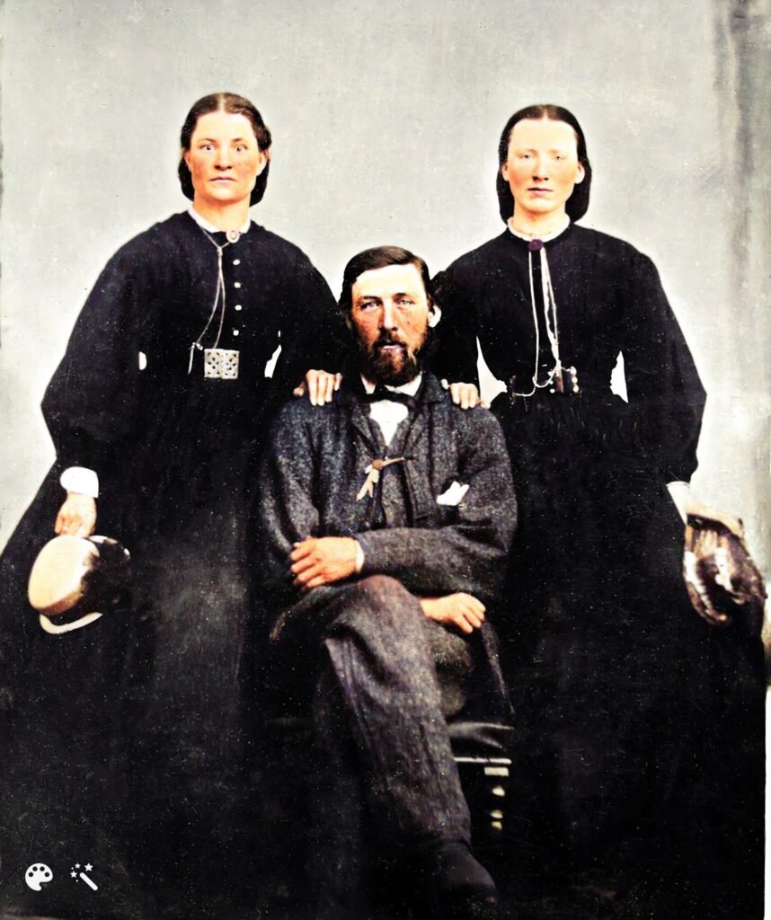 Two women standing with their hands on a sitting gentleman's shoulder