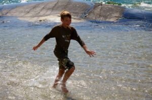 Boy skipping through shallow water with rocks in the background.
