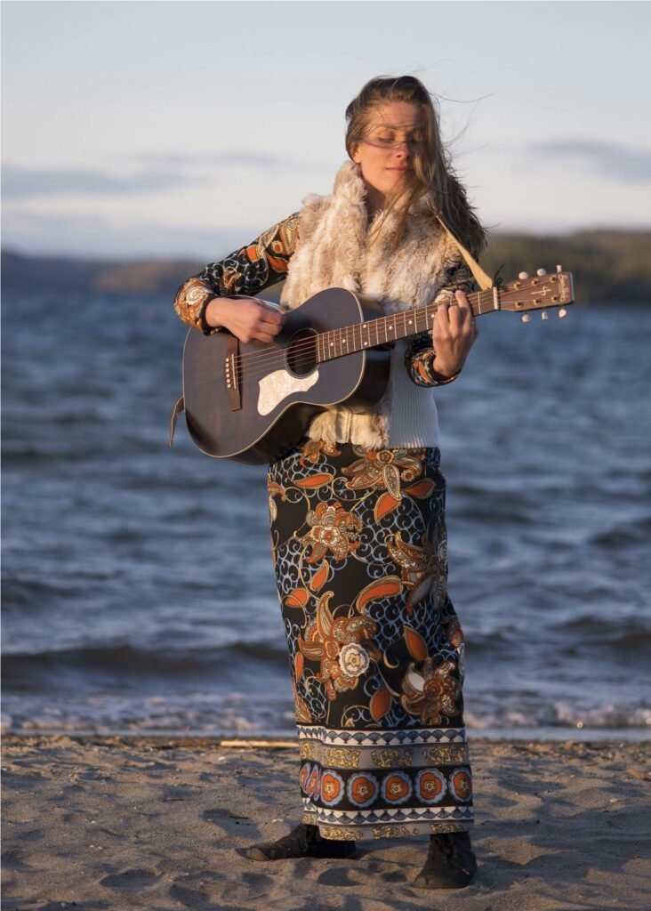a YOUNG LADY STANDING WITH A GIUTAR IN HER HANDS , STANDING WITH A LAKE BEHIND HER.