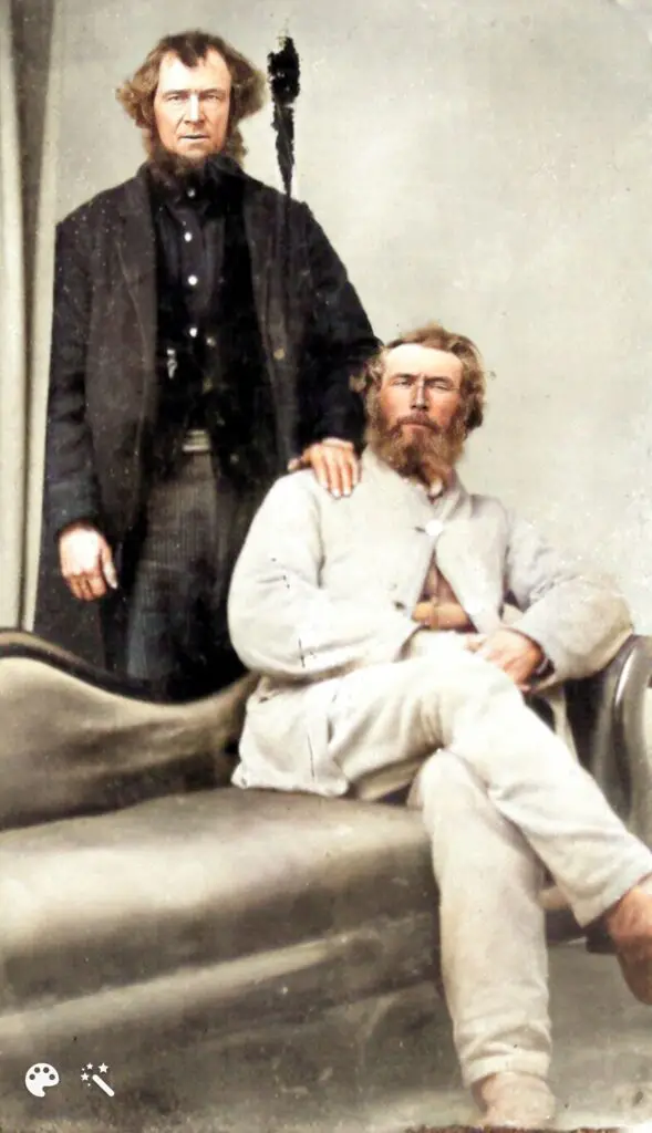 Gentleman in a white suit sitting on a divan with a man behind him with his hand on the sitting man's shoulder.
